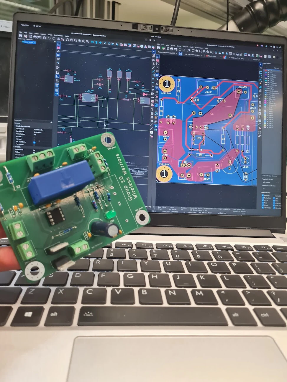 The fabricated green boost converter board held up in front of a laptop showing its KiCad schematic and layout