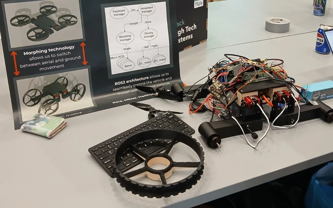 The prototype on a table: a black 3D-printed body with red servos and a nest of wiring, two legs ending in motors, a printed wheel with a pale bearing ring lying in front on a keyboard, and the team poster behind