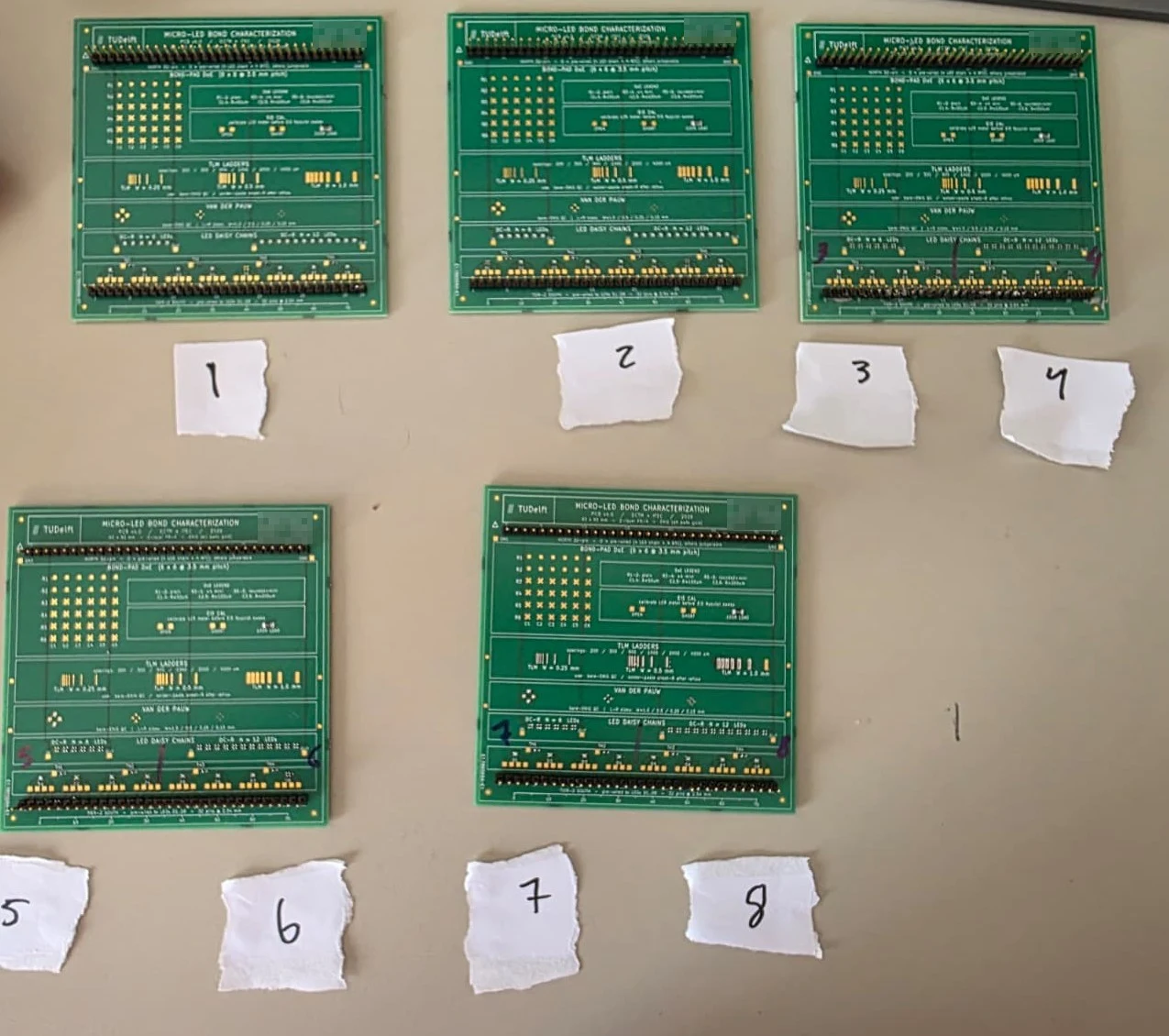 Five fabricated green test boards on a desk with handwritten paper tags numbered 1 to 8 beside them