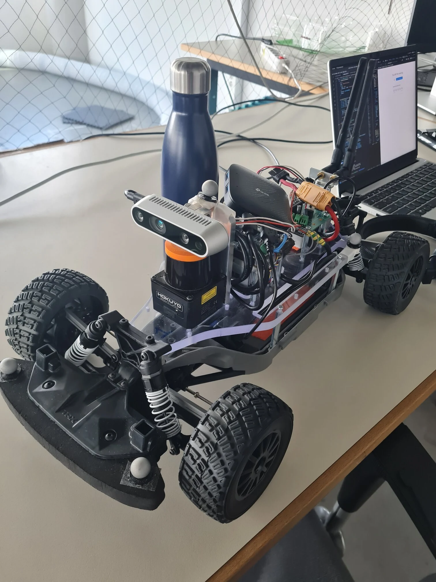 Close view of the F1tenth car on a desk: the black and orange Hokuyo LIDAR at the front under a depth camera, with the onboard computer, wiring and a laptop behind