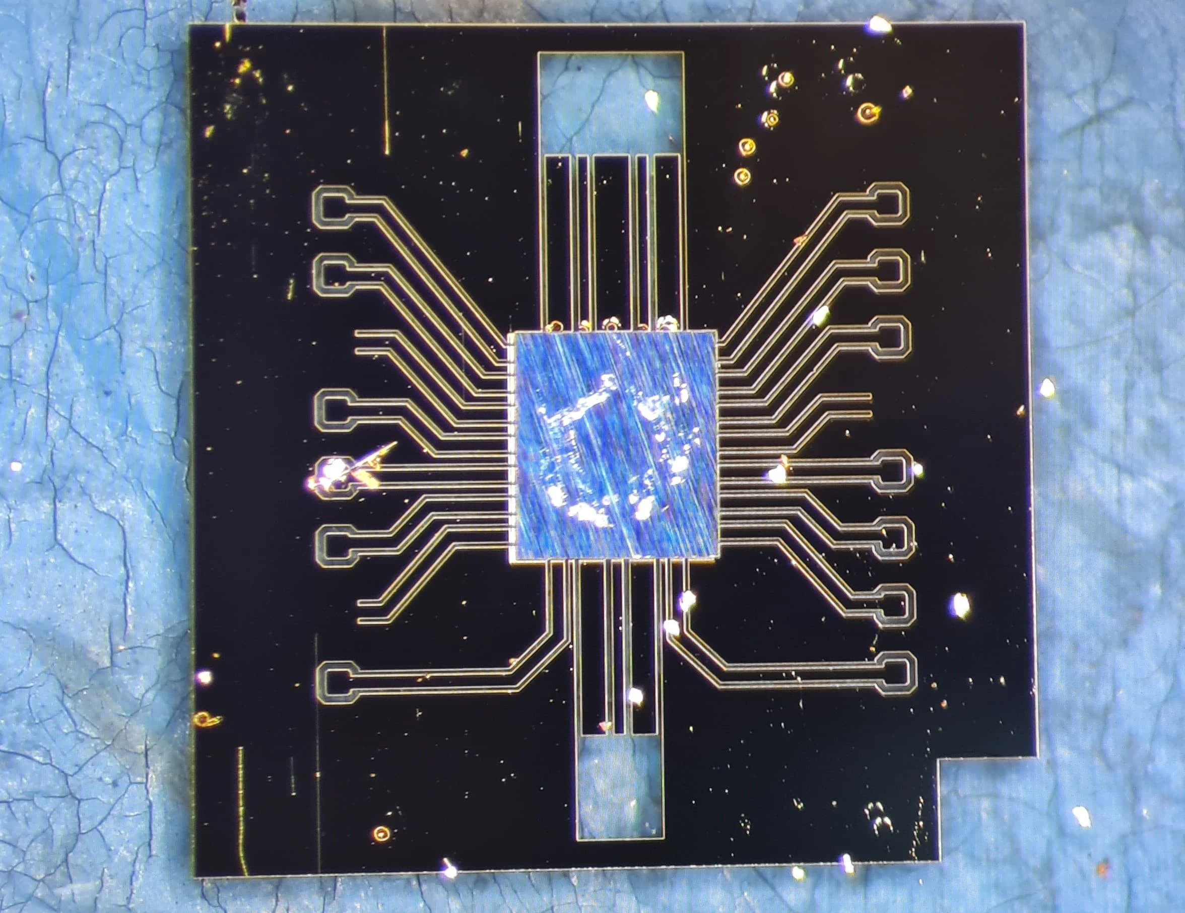 Micrograph of a blue-backed GaN die bonded face-down in the centre of a glass interposer, with gold-outlined traces fanning out to pads on a black background