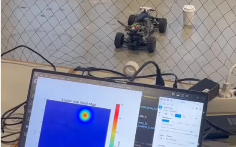 A small LIDAR-equipped RC car on the floor of a netted test area, with a laptop in front showing a blue risk field map with one red and yellow hot spot