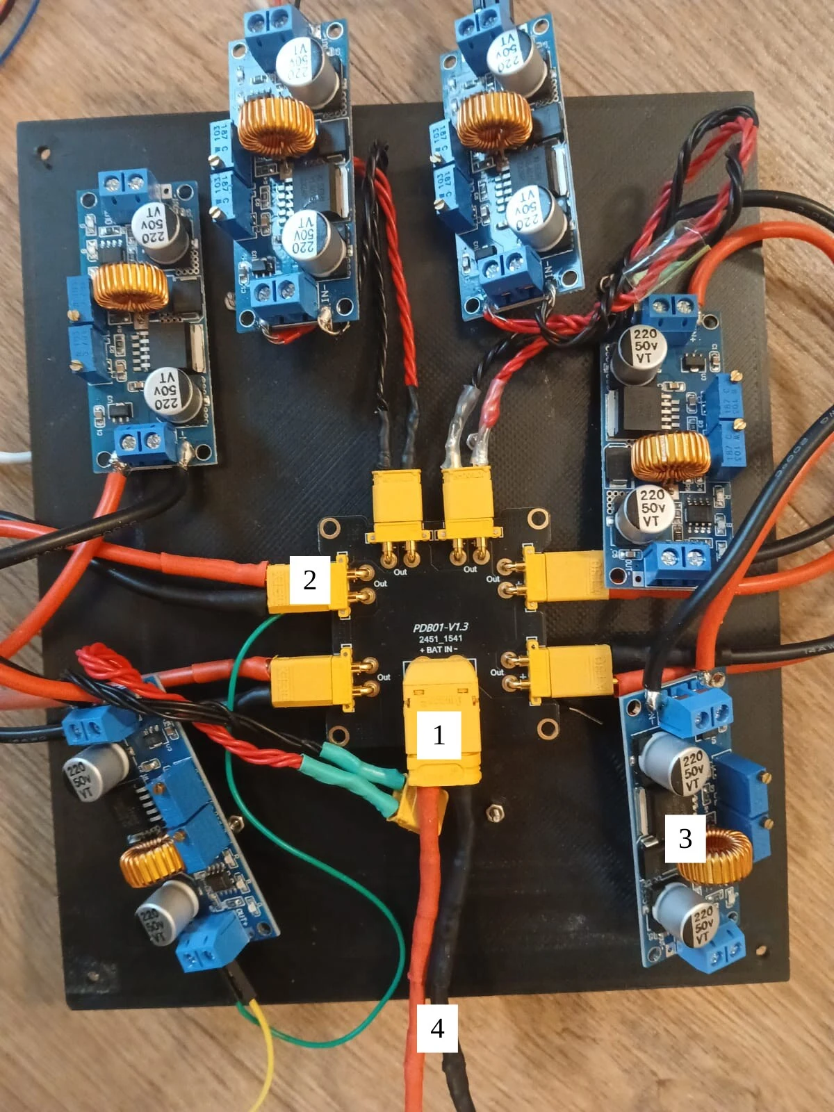 Overhead photo of a black plate with a small power distribution board, yellow XT60 and XT30 connectors and blue buck converter modules wired around it, marked 1 to 4