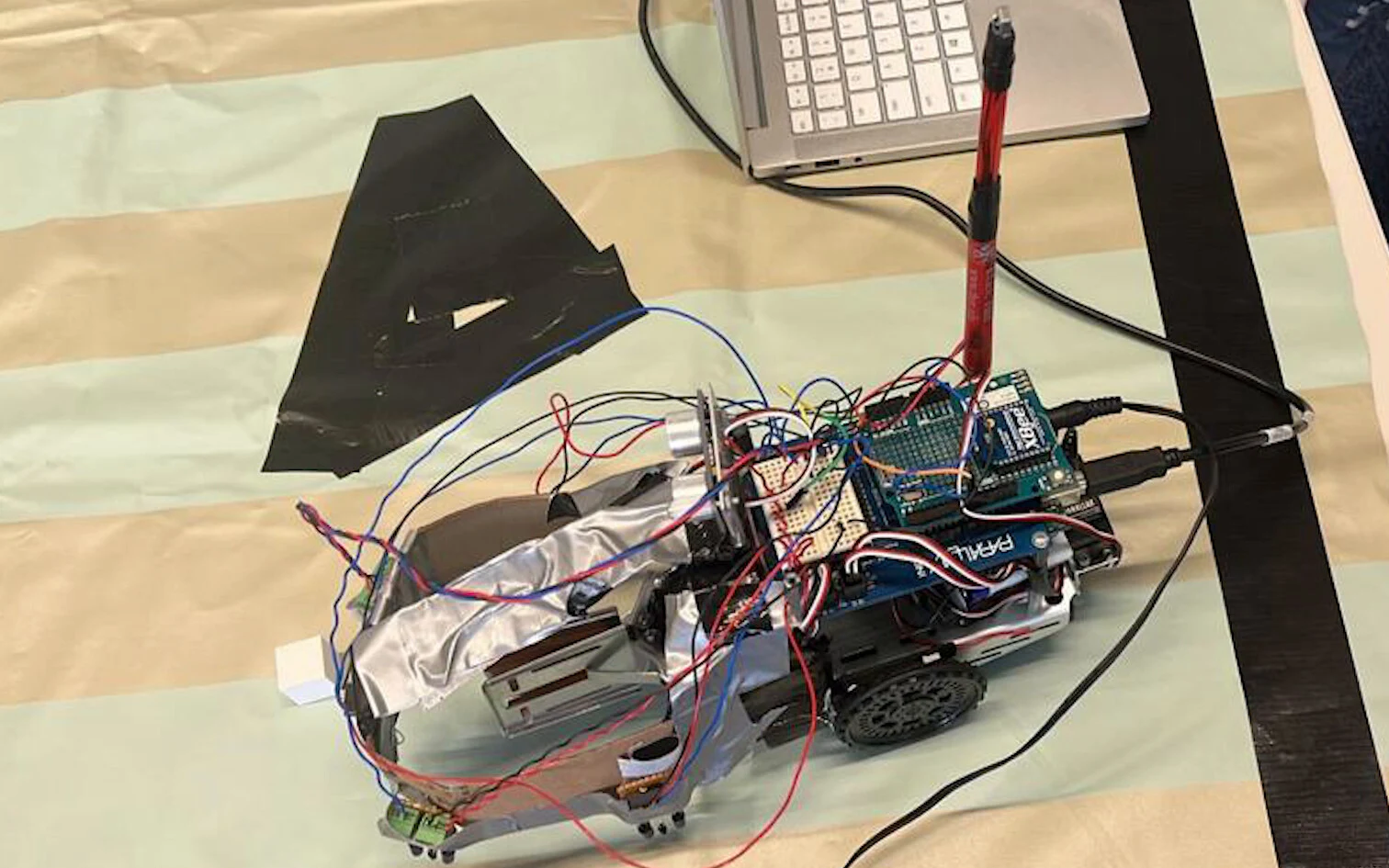 Top-down view of the wired-up rover on the striped practice arena, with its red receiver mast, servo-mounted ultrasonic sensor, gripper and cardboard bumper, beside a patch of black tape